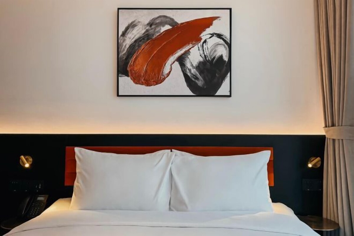 A hotel room with a bed, white pillows, a red headboard, and a framed abstract painting on the wall.