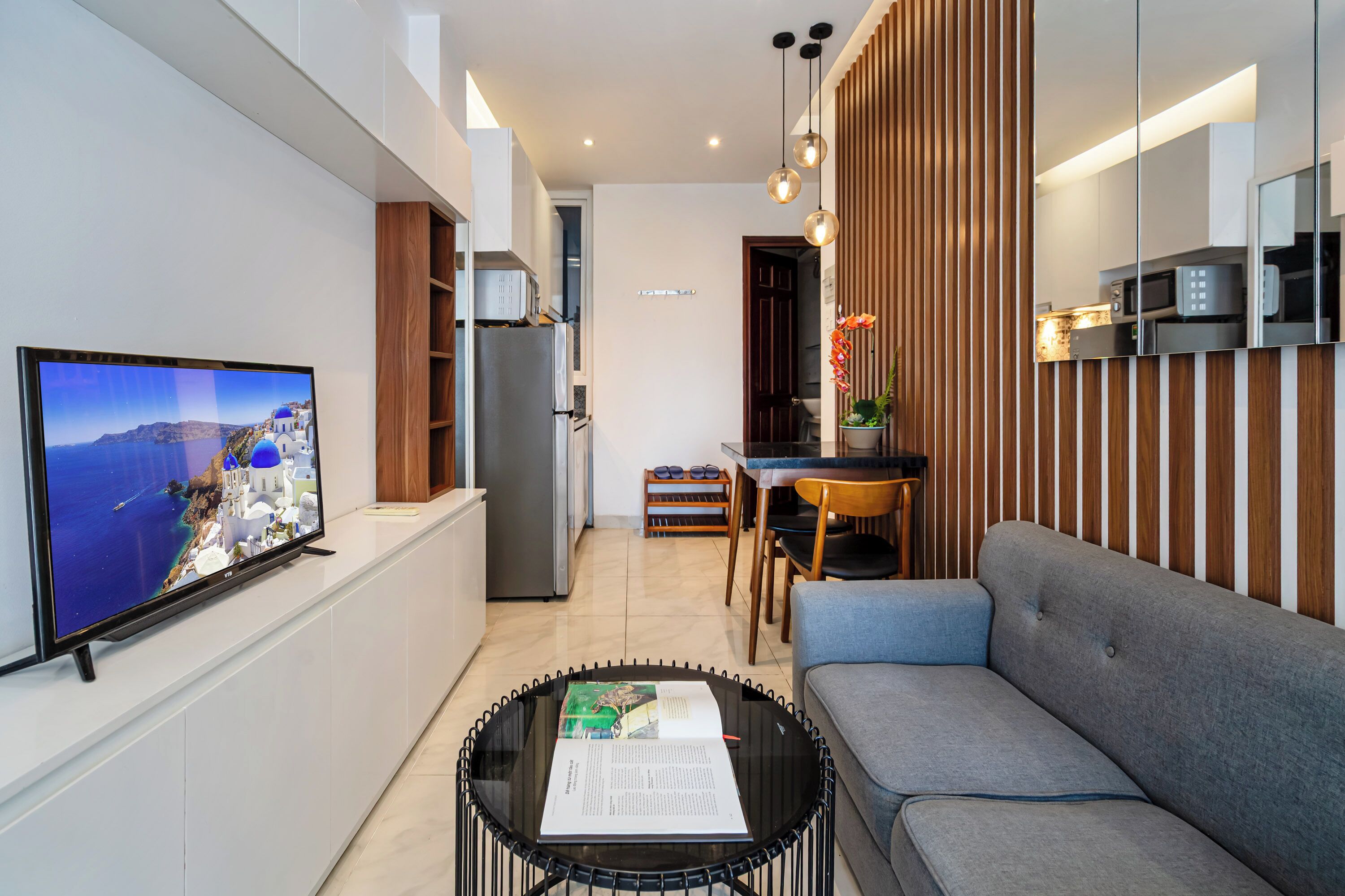 Un salon moderne avec un téléviseur à écran plat, un canapé, une table basse avec un livre, et un coin cuisine à l’arrière-plan.