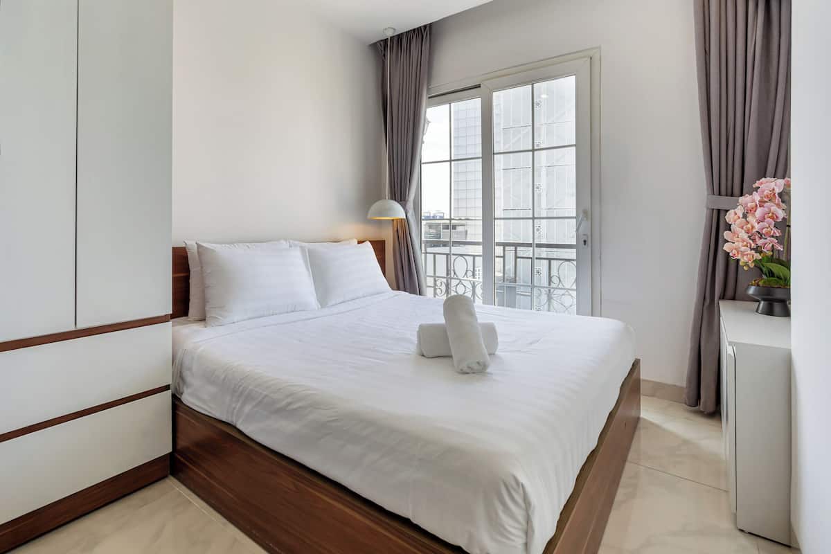 Habitación de hotel con cama, mesita de noche, lámpara, ventana con cortinas y vistas al exterior.