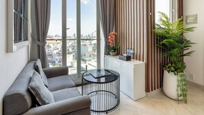 A modern living room with a grey sofa, a glass coffee table, a potted plant, and a view of the city through large windows.