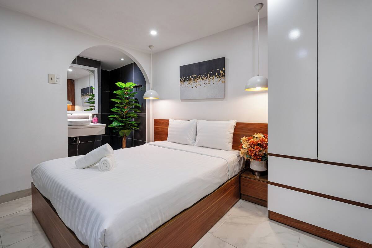 Un dormitorio moderno con una cama grande, una mesita de noche con un florero y un espejo sobre el lavabo.