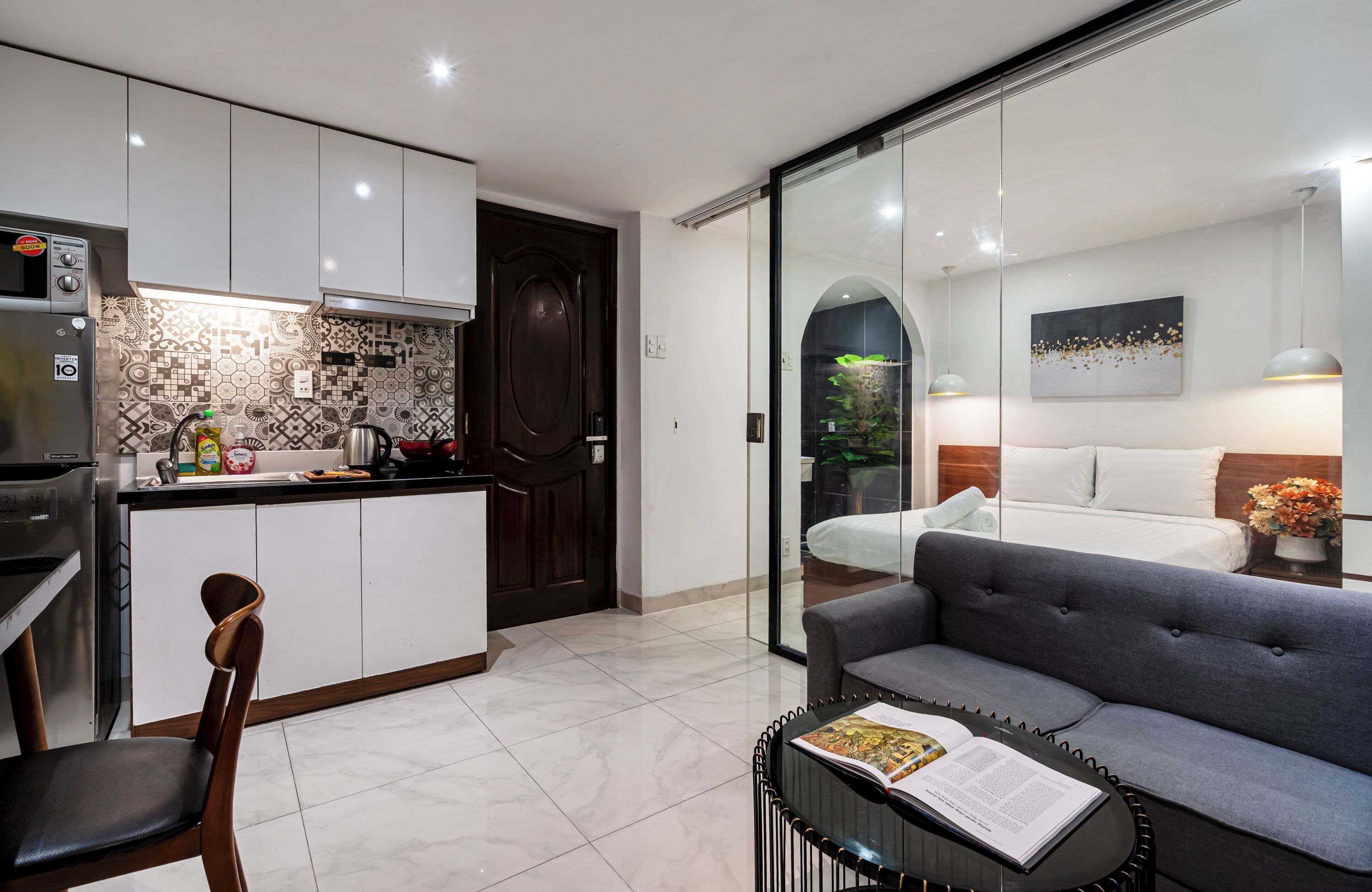 Una cocina moderna con armarios blancos, horno empotrado y salpicadero decorado. Hay un comedor con una silla de madera y una mesa, y una sala de estar con un sofá y una revista sobre la mesa.