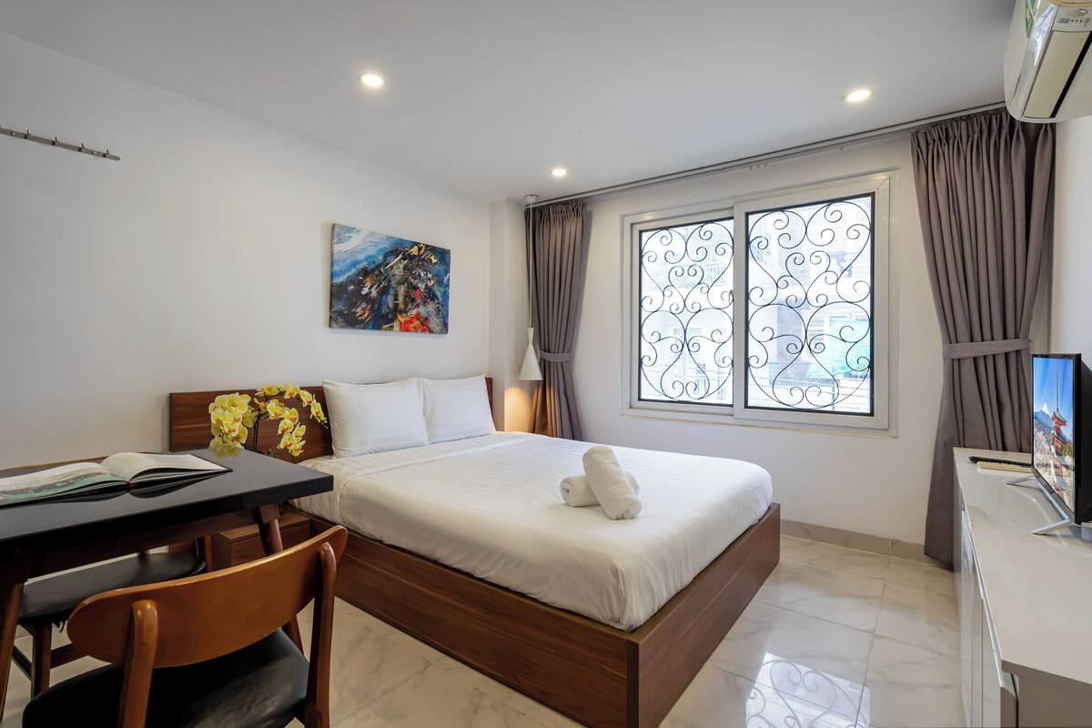 Una habitación de hotel con cama, escritorio con silla, ventana con cortinas, un cuadro en la pared y televisor.