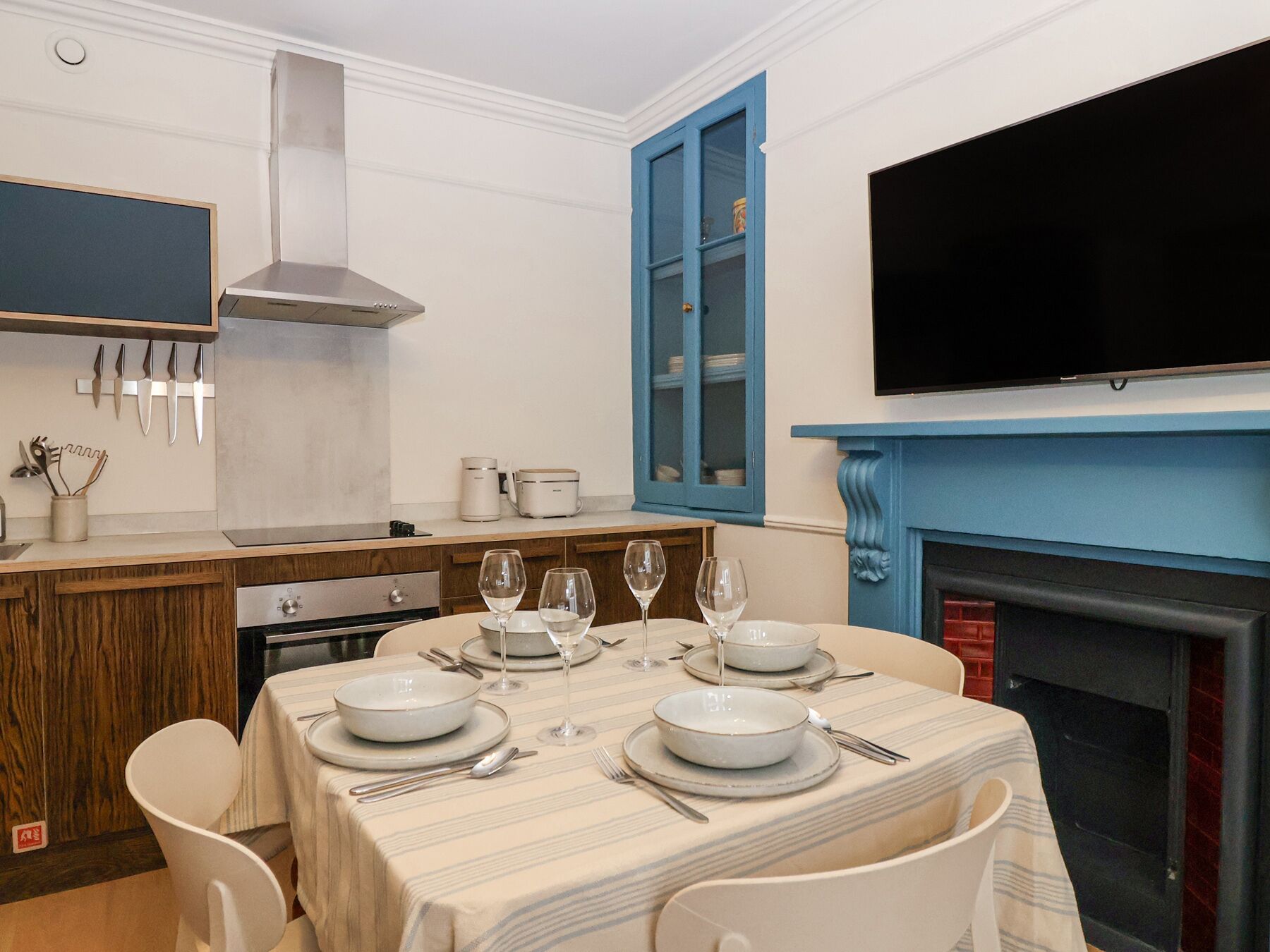 Un comedor con una mesa preparada para cuatro, una cocina con electrodomésticos modernos y un armario azul con puertas de cristal.