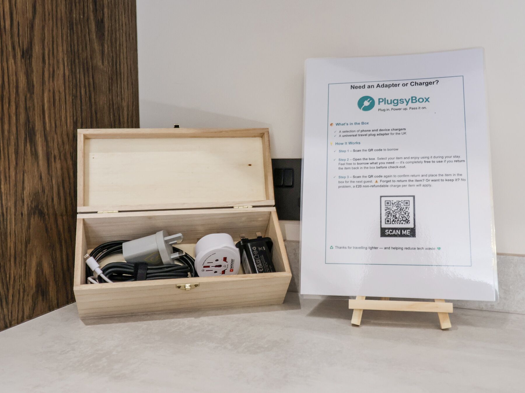 A PluggyBox with a QR code and a wooden box containing various travel adapters and chargers.
