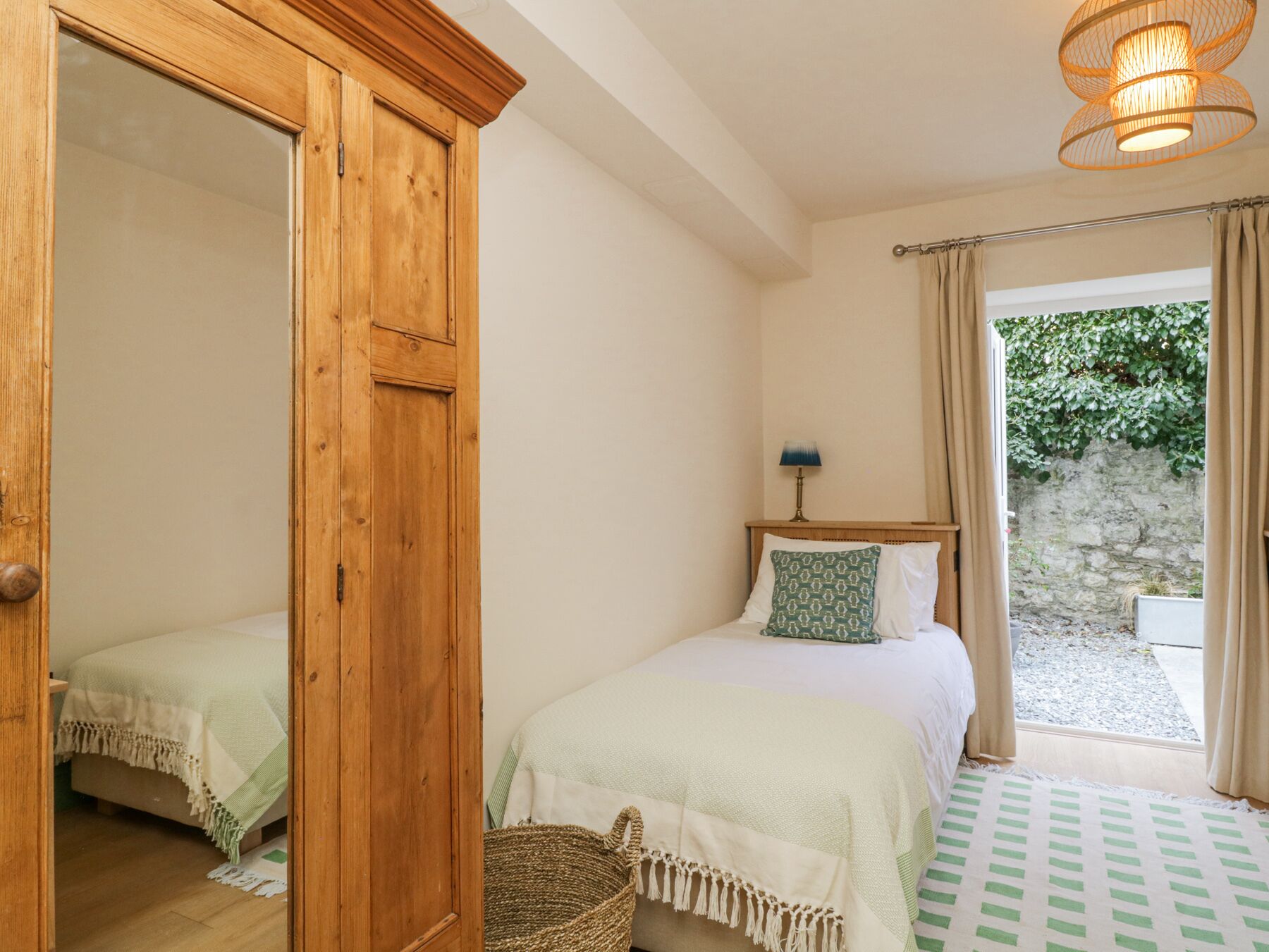 A bedroom with a wooden wardrobe, a bed with a green and white checkered bedspread, a bedside lamp, and a view of a garden through a window.