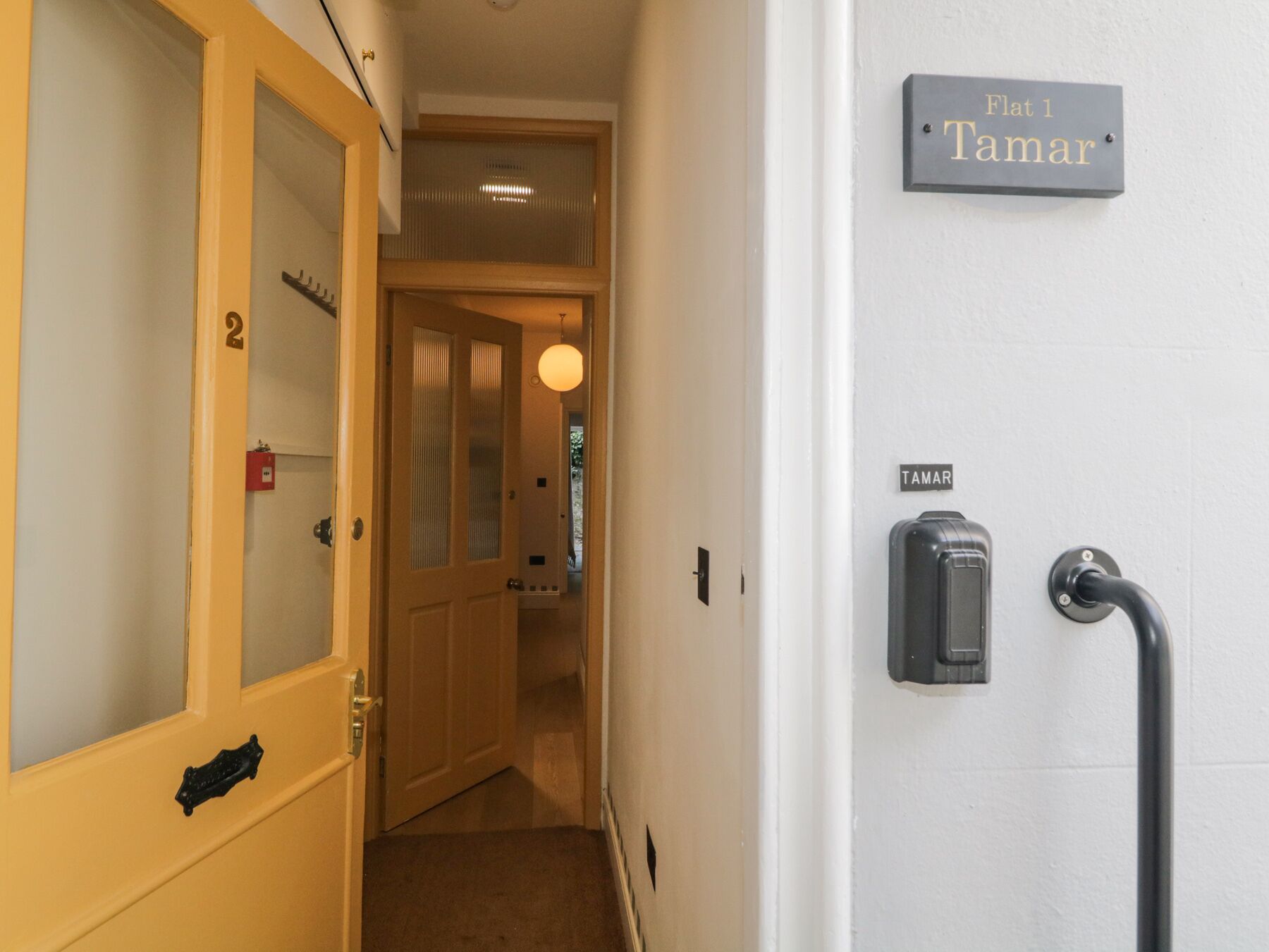 A hallway with a door labeled "Flat 1 Tamar" and a wall-mounted device, possibly a security system.