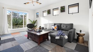 A living room with a gray sofa, a wooden coffee table, and a ceiling fan. There are windows with curtains, a plant, and framed pictures on the wall. - 4Beds Private Unit Near BeachWalk Golf (Pacifica)