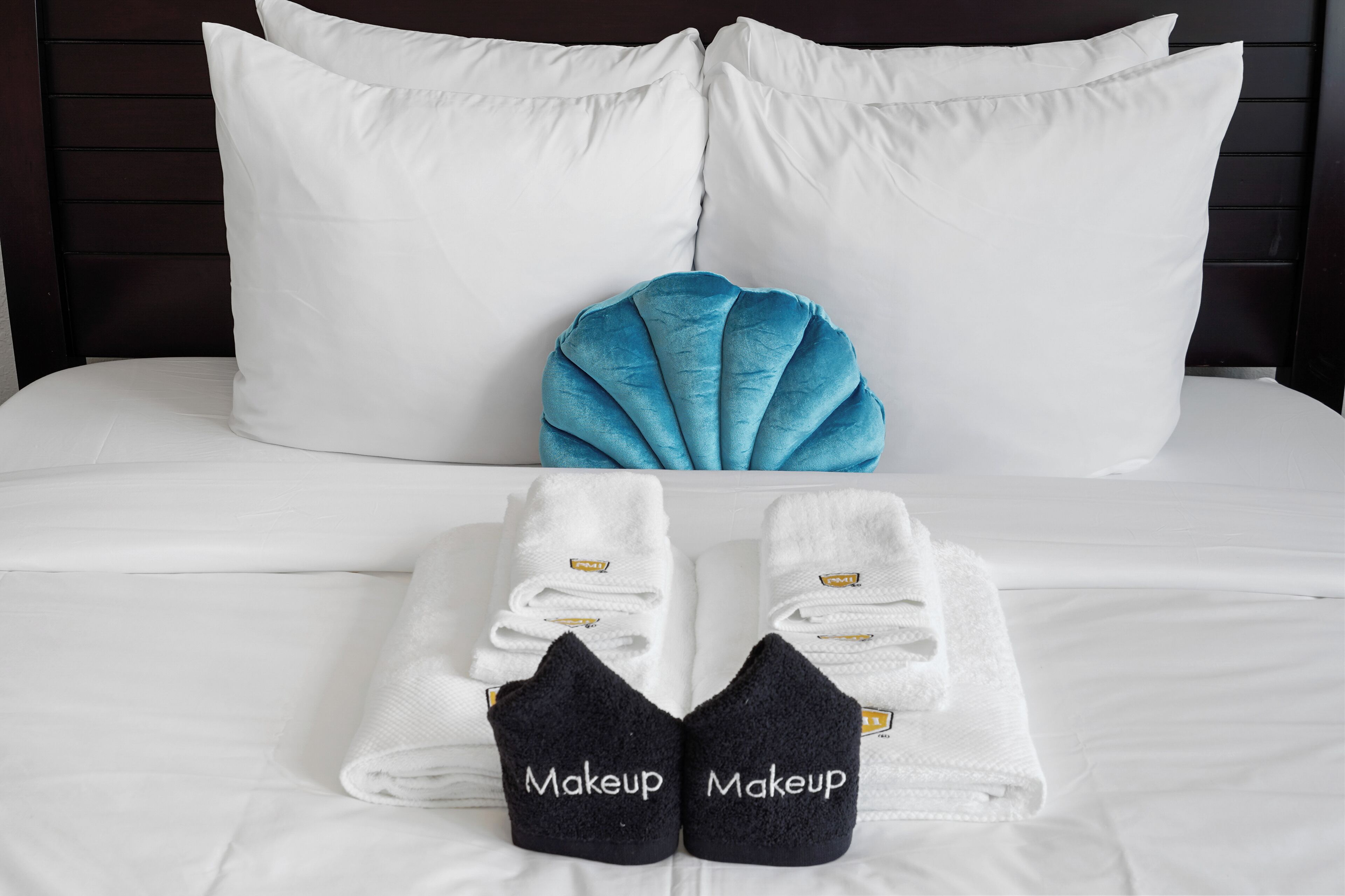 Un lit bien fait, avec des draps blancs, un coussin décoratif bleu et deux serviettes sur lesquelles est écrit le mot « Maquillage ».
