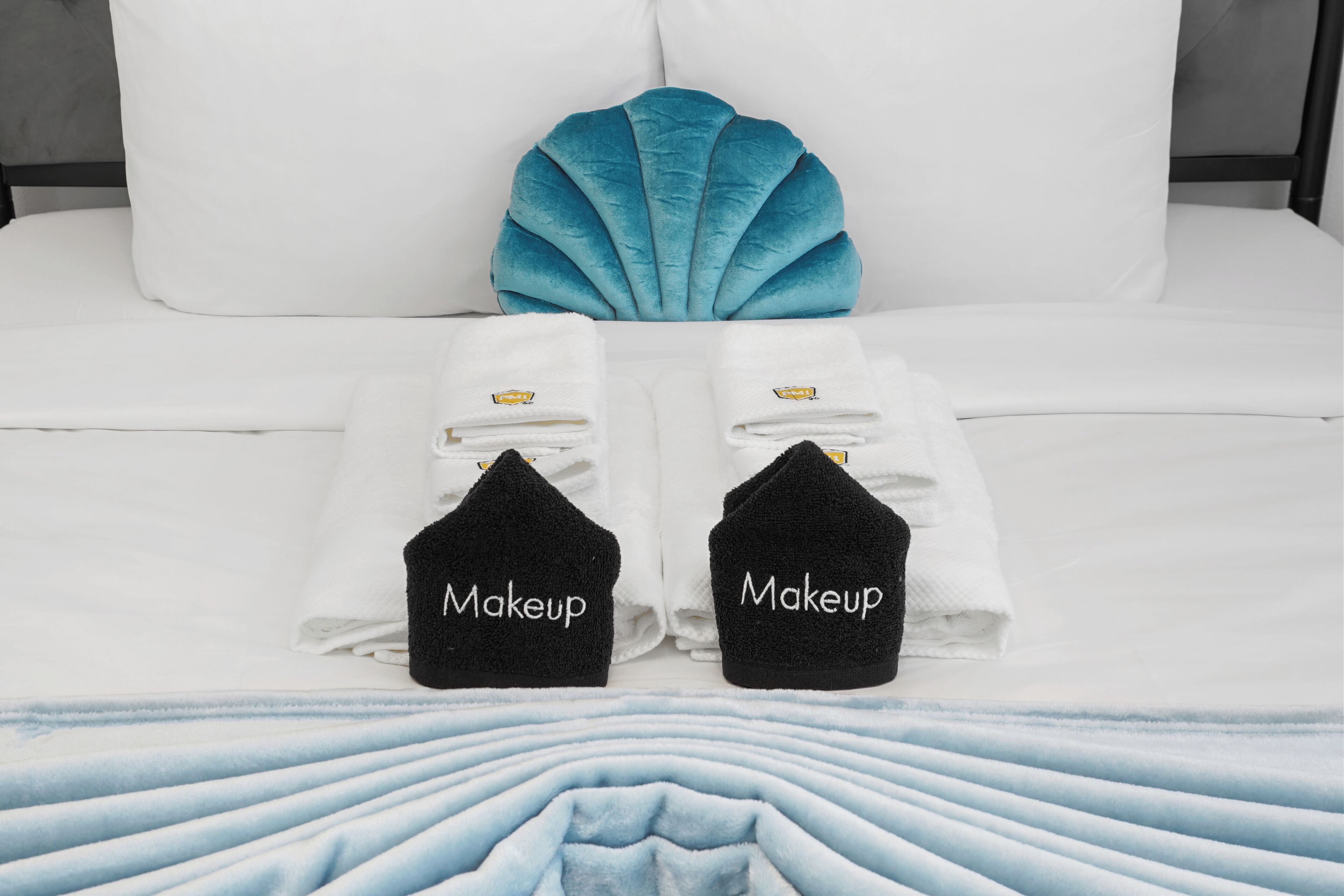 A neatly made bed with a blue decorative pillow, two towels labeled 'Makeup', and a blue patterned blanket.