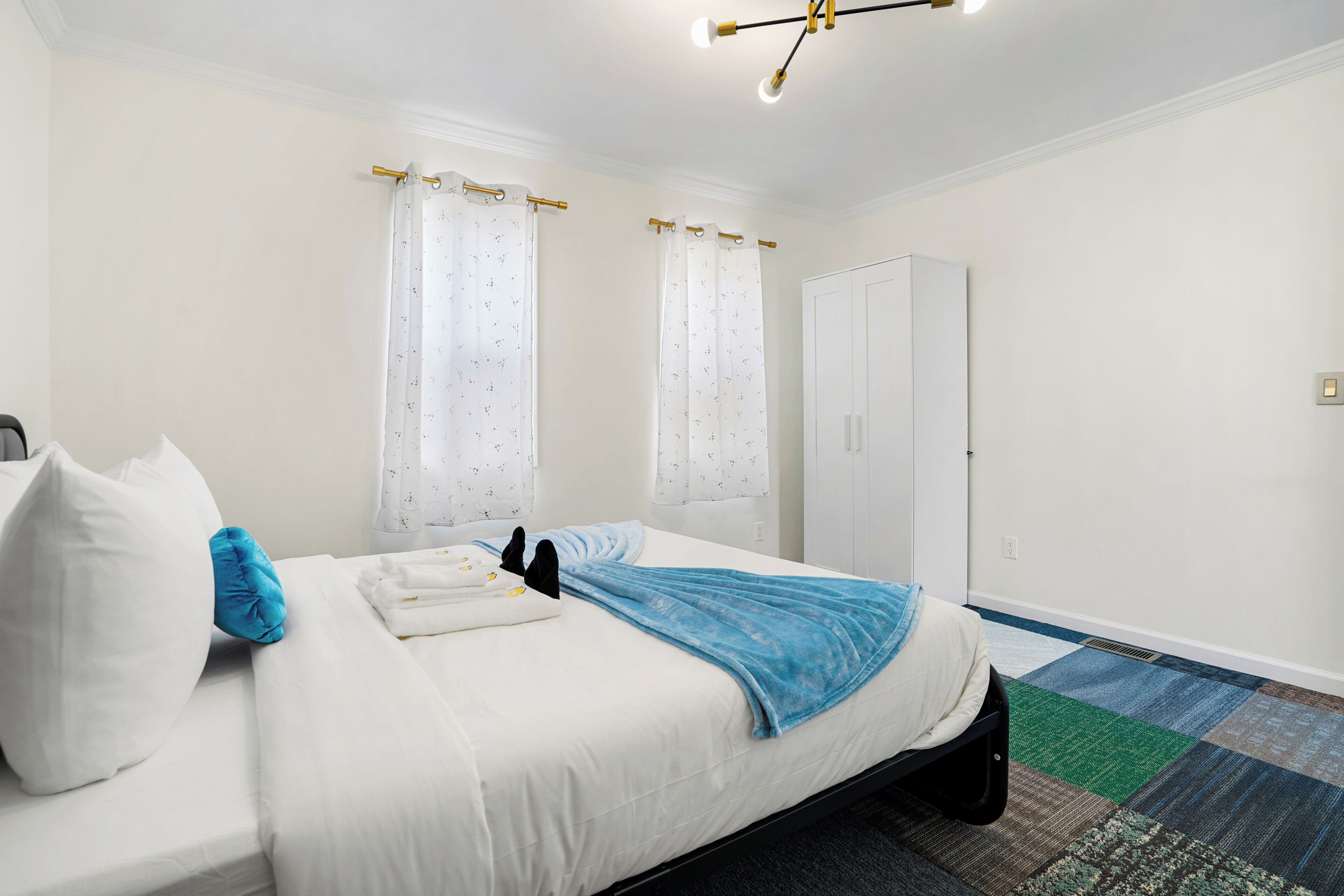 A neatly made bed with white bedding and a blue throw blanket, a white wardrobe, and a patterned carpet.