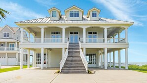 Une grande maison de deux étages située en bord de mer, dotée d’un porche avant couvert, d’un escalier menant à l’entrée et offrant une vue sur l’océan.