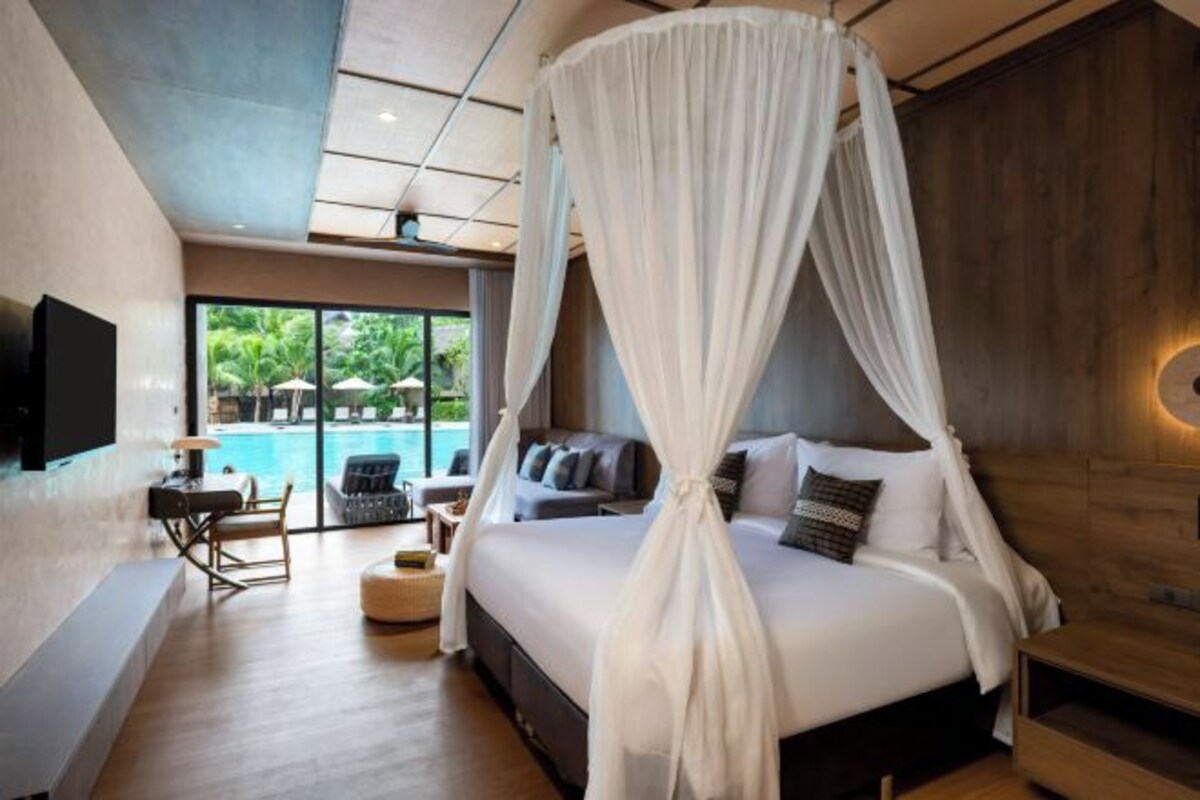 A bedroom with a four-poster bed, a TV, a seating area with a table and chairs, and a view of an outdoor pool.