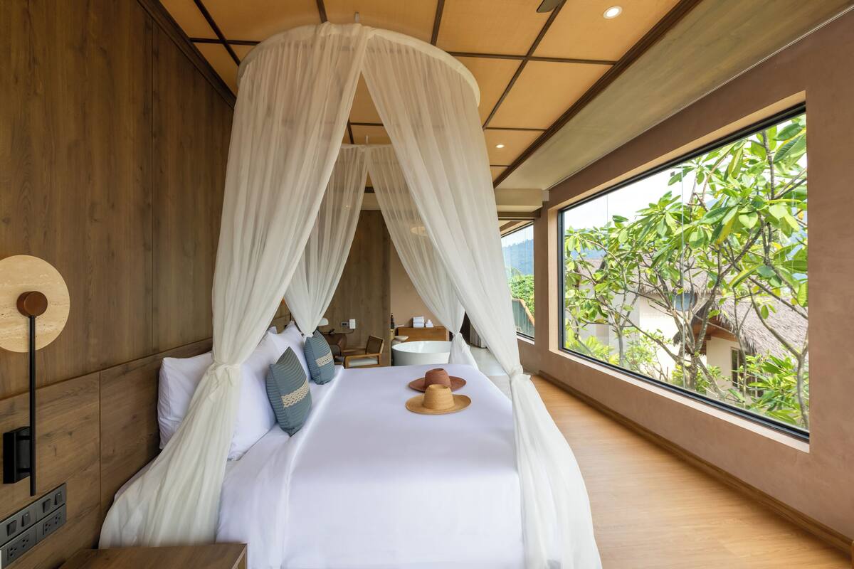 A bedroom with a canopy bed, wooden walls, a bedside table, and a view of greenery outside.