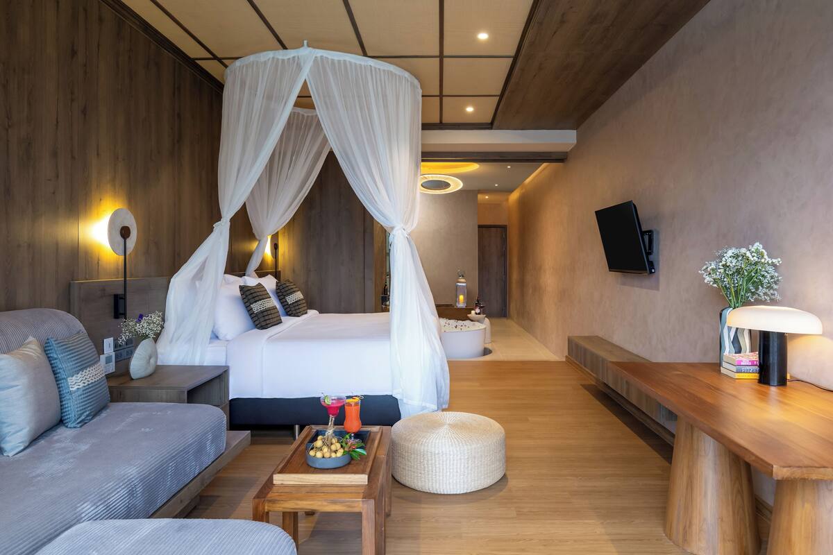A modern bedroom with a canopy bed, a sofa, a bedside table with a tray of fruit and a drink, and a bedside lamp.