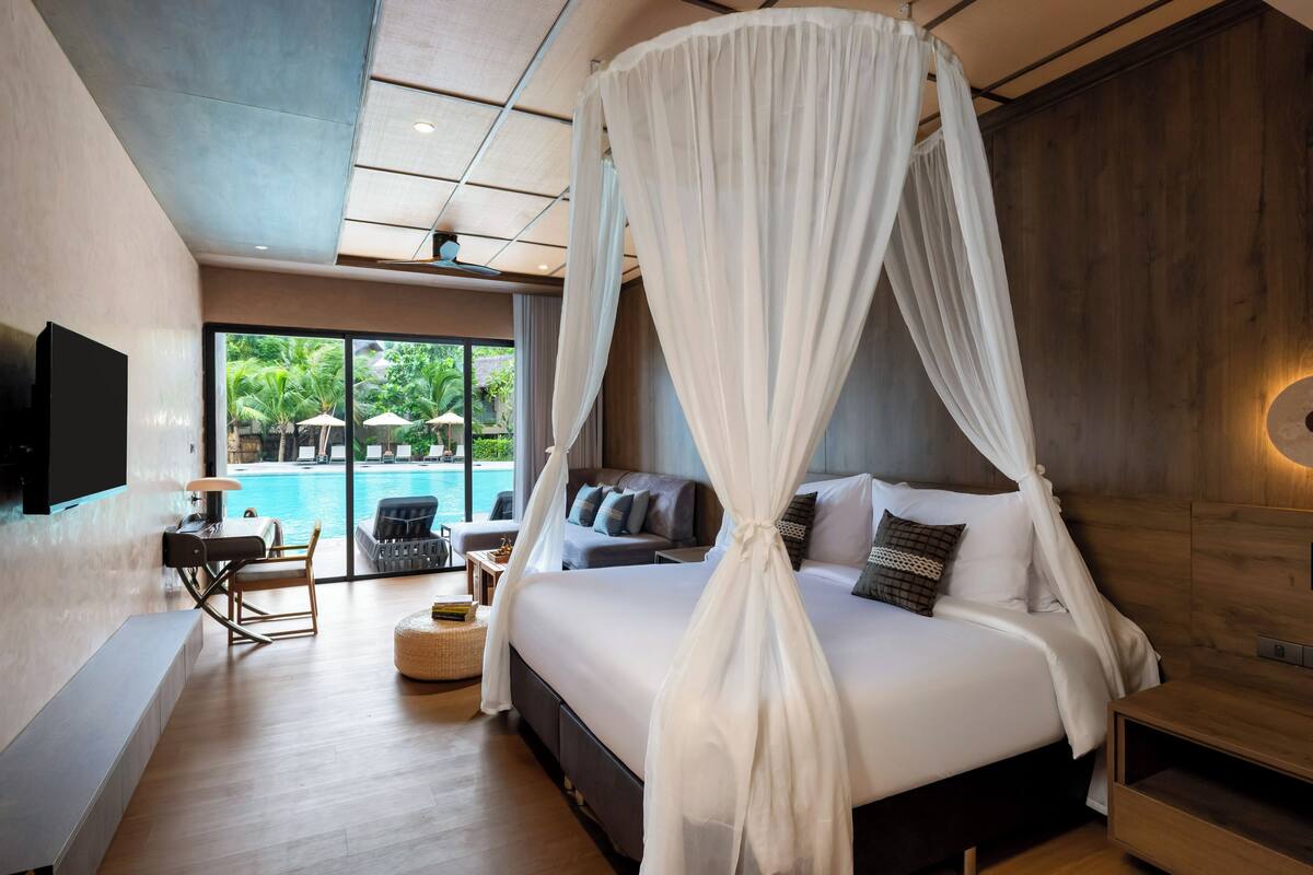 A bedroom with a four-poster bed, a TV, a seating area with a table and chairs, and a view of a swimming pool.