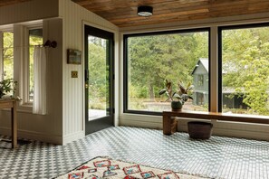 A room with a checkered floor, a large window with a view of a house and trees, a wooden ceiling, and a door with a coat rack. - The Carly Fern Lodge | Luxe Wine Country Experience, Entire Lake, Trails & 21 Gorgeous Acres (Carlton)