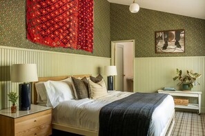 Ein Schlafzimmer mit einem Bett, zwei Nachttischen mit Lampen, einem Spiegel, einer Wand mit grünem Muster und einem roten, hängenden Wandbehang.