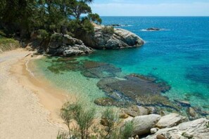 Paisagem costeira com águas turquesa cristalinas, praia arenosa, afloramentos rochosos e vegetação densa.