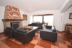 Uma sala de estar com lareira de pedra, sofás de couro, área de refeições com cadeiras e vista através de amplas janelas.