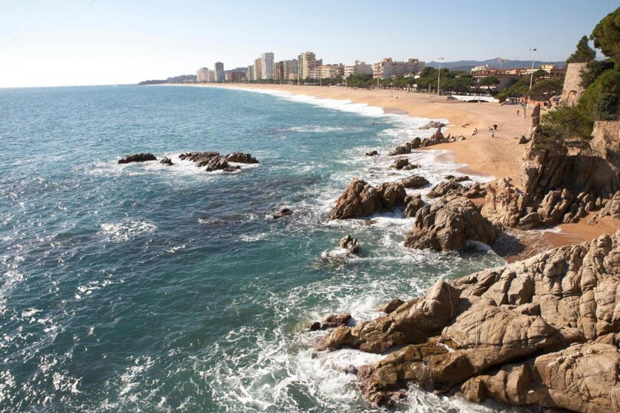 Un paisaje urbano costero con un litoral rocoso, una playa arenosa y un horizonte de edificios al fondo.