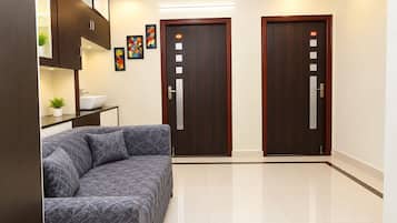 Una habitación de hotel moderna con sofá, una mesita con un frutero y una planta, dos puertas de madera con paneles numerados y suelo de baldosas.