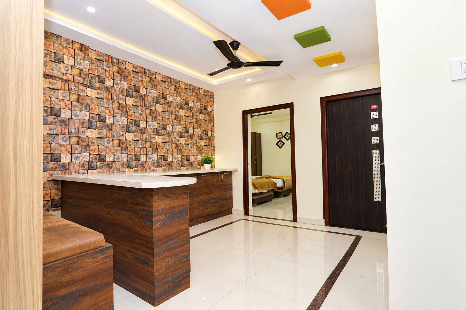 Zona de recepción de hotel con un mostrador de madera, una pared con diseño de mosaico, un ventilador de techo y una puerta con panel de seguridad.