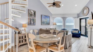 A spacious living area with a round glass table, white chairs, a sofa, and a staircase with wooden railings. - 518 Seascape | Waterfront Views + Community Pool (Isle Of Palms)