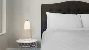 Una cama bien hecha con sábanas blancas, una mesita redonda con una lámpara y un cabecero tapizado oscuro.