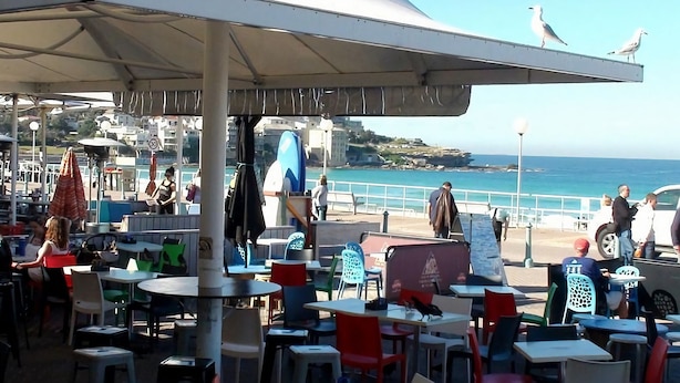 Un café al aire libre con mesas y sillas, con vistas a la costa y al mar, y edificios a lo lejos.