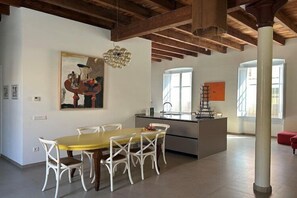 A spacious dining area with a yellow table and white chairs, a large painting on the wall, and a modern kitchen with a window.