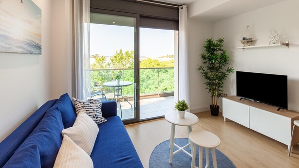 Una sala de estar moderna con un sofá azul, un televisor de pantalla plana, una pequeña mesa redonda y un balcón con vistas a zonas verdes.