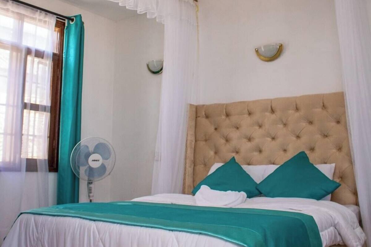A bedroom with a tufted headboard, a bed with white and teal bedding, a teal curtain, a white fan, and a window with a curtain.