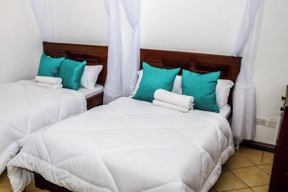 Two twin beds with white bedding and teal pillows, a wooden headboard, and a white curtain.