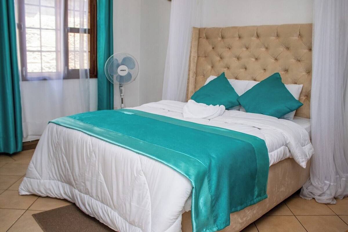 A neatly made bed with a teal bedspread and white bedding, a tufted headboard, and a standing fan beside the window.