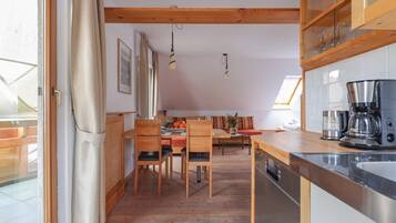 Eine moderne Küche mit Holzsäulen, eine Essecke mit Holztisch und -stühlen sowie ein Blick ins Freie durch eine Glastür.