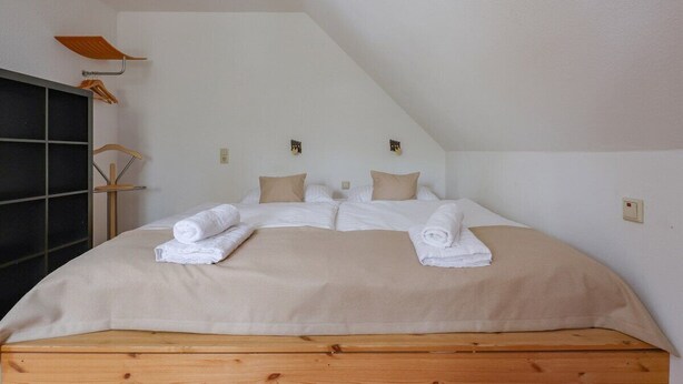 A neatly made bed with a beige bedspread and pillows, a wooden headboard, and a small shelf with towels. - 4 person apartment in green suroundings (Berlin)