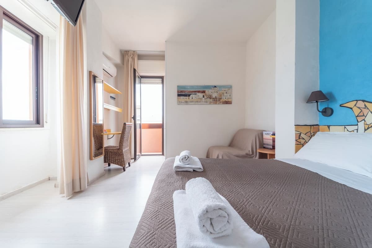 A hotel room with a bed, a chair, a desk, and a painting on the wall.