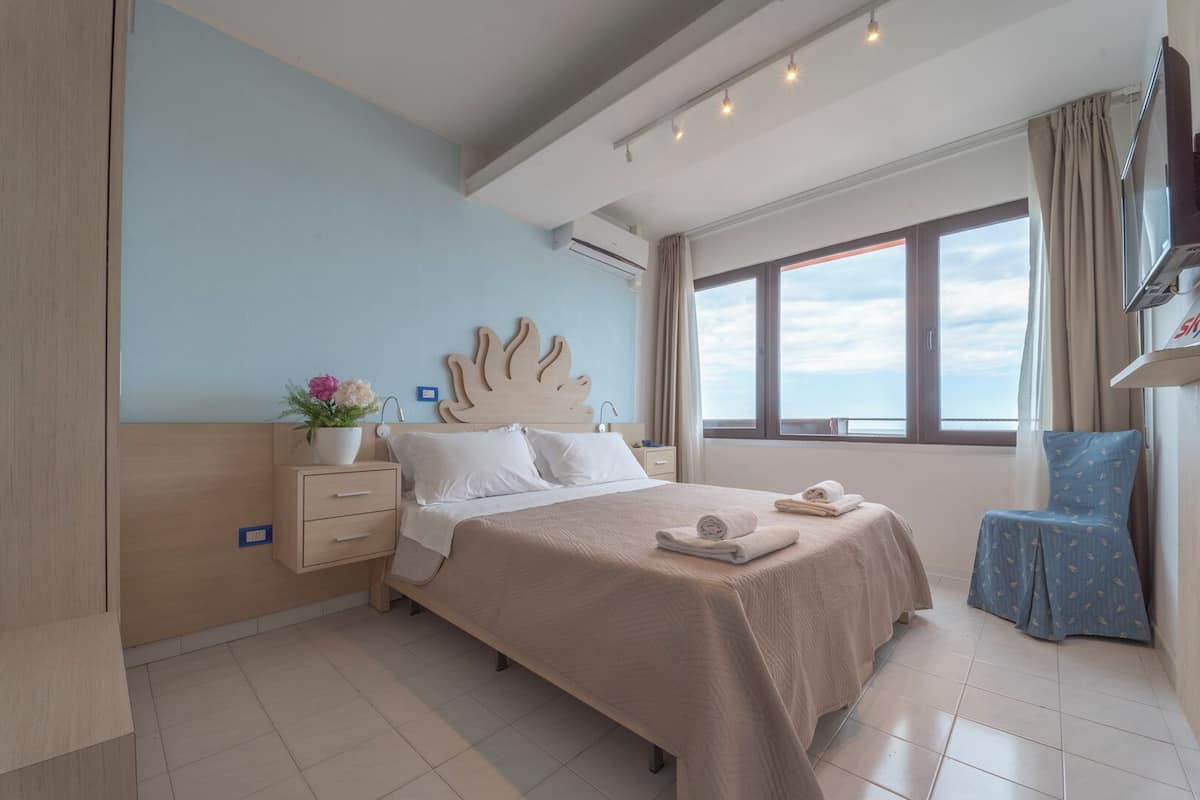 A modern hotel room with a large bed, a bedside table with a lamp, a small chair, and a view of the sea through the window.