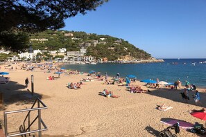 Una playa con gente tomando el sol y nadando, cielo despejado y edificios en una colina al fondo.