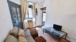 A living room with a sofa, ottomans, a glass coffee table, a television, a cabinet with glass doors, a chair, and a window with curtains.