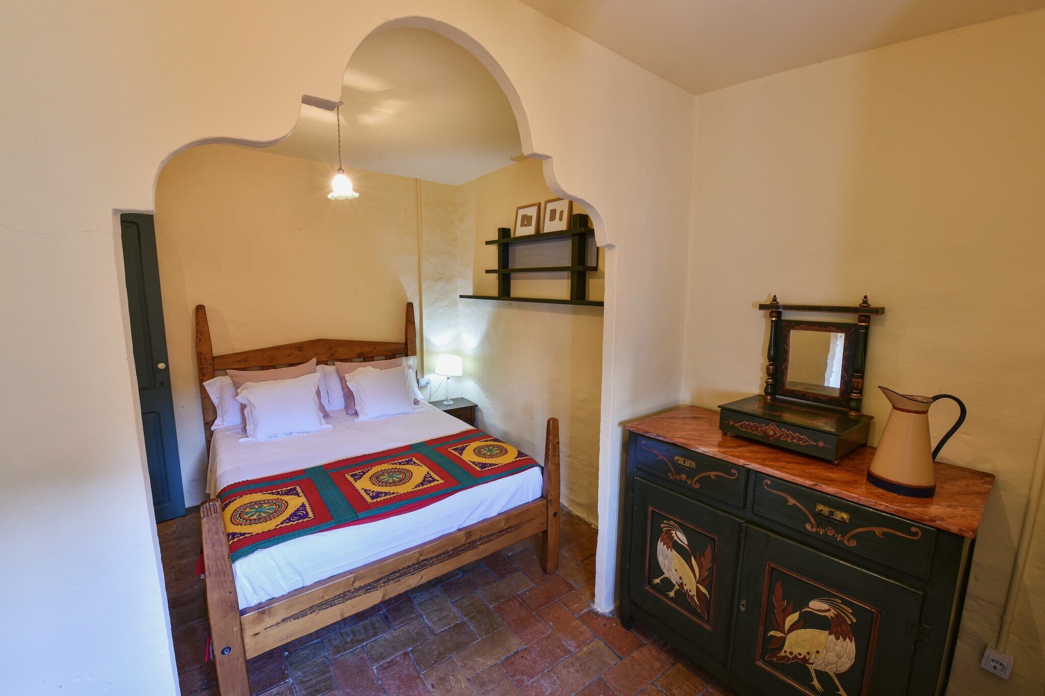 A bedroom with a wooden bed, a nightstand, a dresser with a mirror, and a shelf with framed pictures.
