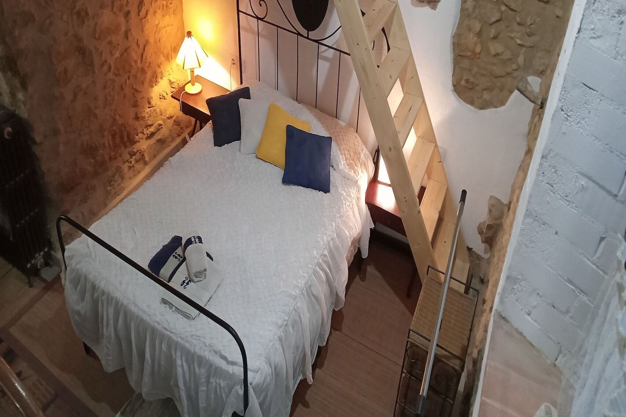 A bedroom with a stone wall, a wooden ladder, a bed with white bedding and pillows, and a nightstand with a lamp.