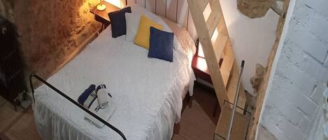 A bedroom with a stone wall, a wooden ladder, a bed with white bedding and pillows, and a nightstand with a lamp.