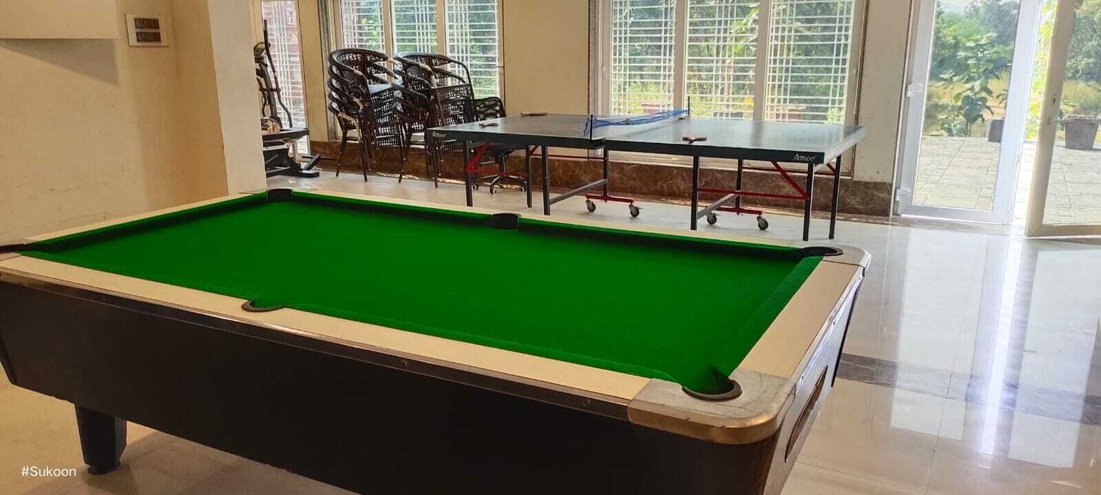 A pool table with a green felt surface, a seating area with chairs, and a view of greenery through large windows.