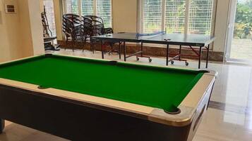 A pool table with a green felt surface, a seating area with chairs, and a view of greenery through large windows.