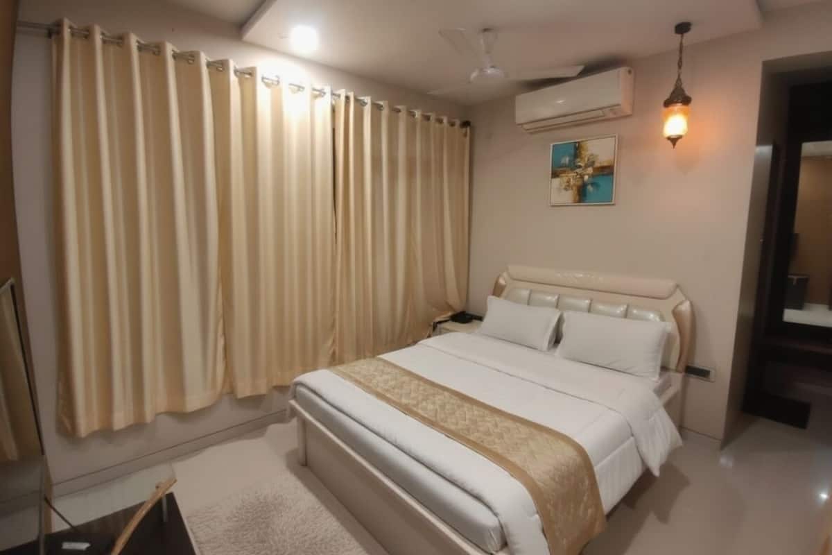 Quarto de hotel com uma cama grande, cortinas bege, ventilador de teto, ar condicionado e mesa de cabeceira com abajur.
