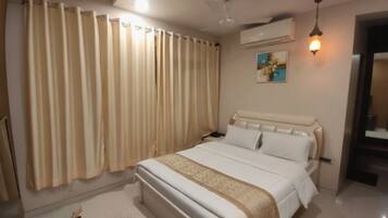A hotel room with a large bed, beige curtains, a ceiling fan, an air conditioning unit, and a bedside table with a lamp.