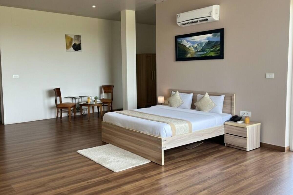 Quarto moderno com piso de madeira, cama com roupa de cama branca, ventilador de teto, aparelho de ar condicionado e um quadro na parede.
