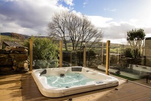 Un jacuzzi en una terraza de madera con recinto de vidrio, con vistas a un paisaje rural con colinas y un árbol.